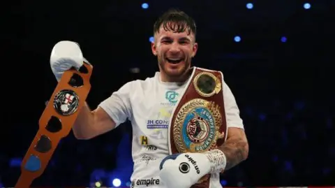 Owen Cooper, a man in a white tshirt with logos on is wearing white boxing gloves and holding two boxing belts