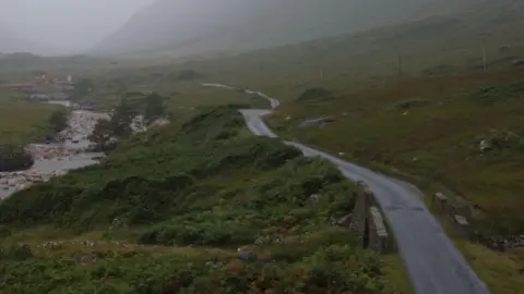 A large green landscape with a river on the left of the image and a road running on the right hand side. The road disappears into the distance and it's a misty day.