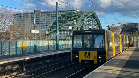 A black and yellow Tyne and Wear Metro train travelling towards Newcastle Airport is approaching St Peter's Metro station in Sunderland. The Wearmouth Bridge is in the background. It is a green archway structure. The Echo Building - a tall, glass building with apartments - is also visible across the bridge. 