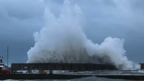 A large wave crashes against a harbour wall at Stonehaven. It is a dark, wet afternoon.
