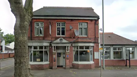 Google The Royal Oak Pub has a large tree on the pavement outside it. it is a red brick pub with grey/green panels around its windows and entrance way.