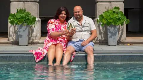 A woman with a pink flowery dress and dark hair sits next to a man who has a shaved head and beard. He is wearing light blue shorts and a light short-sleeved shirt. They are toasting with champagne glasses and their feet are in a swimming pool. 