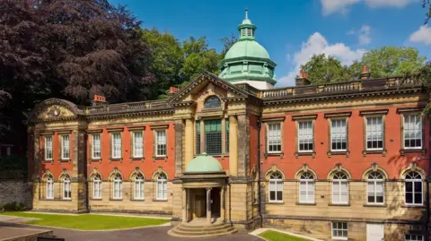 Front view of a historic red-brick and stone building with symmetrical windows, a columned entrance, and a green copper dome, set among mature trees.