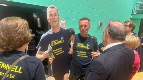 Kieran Haddock/BBC Ed Gemmell in a group of people indoors gathered around a life-size campaign cut-out, with several wearing navy T-shirts reading “Vote Ed Gemmell – Pro Business, Pro Climate, Pro Wycombe,” while others in suits stand nearby in what appears to be a sports hall with green walls and a wooden floor.
