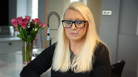 BBC A blonde woman with dark-rimmed glasses, wearing black sits at a kitchen island with pink roses in the background. She looks into the camera with a serious expression on her face.