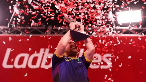 Getty Images Luke Littler is posing with a trophie as confetti rains down around him. He is smiling.