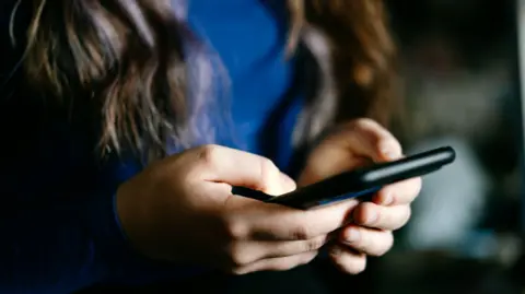 Getty Images A girl with long brown hair and wearing a blue top holds a mobile phone in a black case.