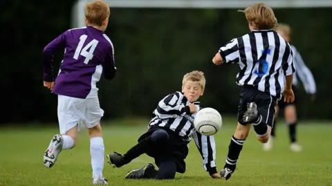 Three boys, playing football. One is on the ground, with a football near to his face, and his eyes closed. Two others have their backs to the camera and are running. Another child is blurred and in the distance. 