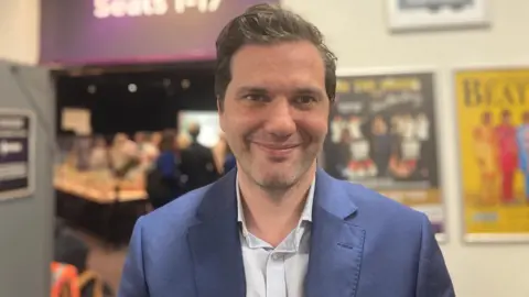 Laura Coffey/BBC Oliver Cooper smiles at the camera standing next to the election count room. He wears a blue suit over a grey shirt.