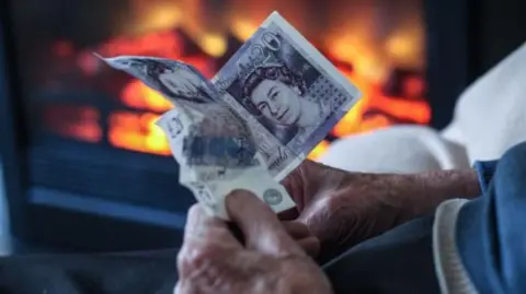 Getty Images an older person's hands grasp two £20 notes in front of a lit fire