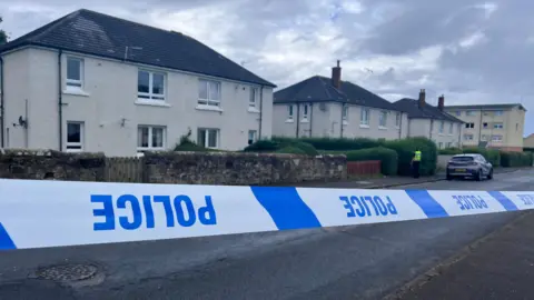 
Blue and white police tape which is upside down in front of a row of cream two-storey buildings. A police officer is visible in a distance.