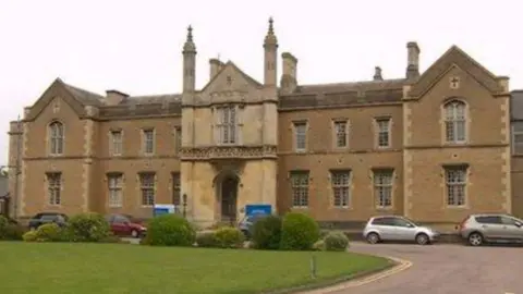 Historic mustard coloured stone building with chimneys and 18 windows and one door. There are cars parked in front of it, some bushes and a grassed area.