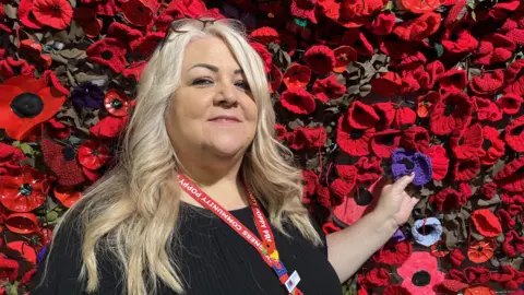BBC Tracy Turner, wearing a black top and a red lanyard, stands in front of a wall of red poppies. She has blonde hair and glasses.