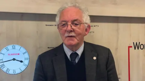 A white haired man with glasses and a white moustache and dressed smartly with a shirt, tie and jacket is standing inside one of the carriages on the exhibit train. He is smiling.