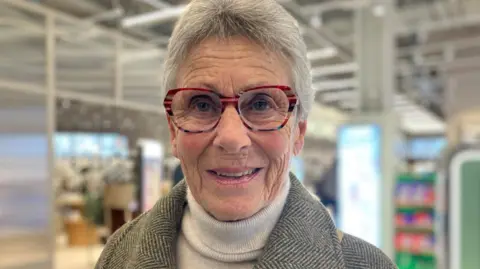 A woman with short grey hair and wearing tortoiseshell reading glasses. She is wearing a beige jumper and a tweed coat. In the background can be seen a retail space
