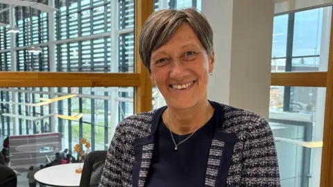 Ben Schofield/BBC Gill Worgan, smiling broadly and looking directly down the camera. She is wearing a blue top, with a white and blue patterned, collared jacket over the top. She has blue eyes and short hair, which is styled in a parting and falls just to her ears. She has two small hooped earrings on and a silver chain hangs around her neck. She is sitting in an office, with large windows behind her, which look out onto an atrium area, in which a handful of people can be seen. 