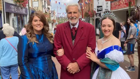 Martin Heath/BBC Grace Magee with long brown hair wearing a petrol blue dress, Lawrence Wheeler with short white hair wearing a long red coat, and Jennifer Taylor-Lee wearing a tiara and a white dress with a blue sash