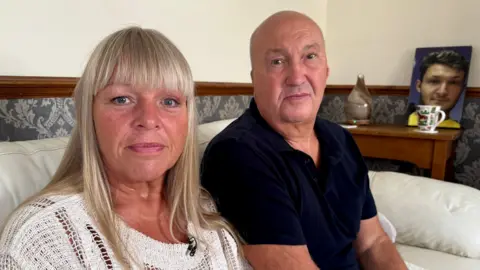 Charlie Rose/BBC Sitting together at home on a white leather sofa, Helen Low, with long blonde hair and wearing a white top, and Martyn Low, bald and wearing a dark blue short-sleeved shirt, look at the camera lens. Behind them is a picture of their son, George.