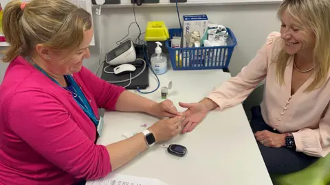 Two individuals seated at a table in a medical setting. One, wearing a pink cardigan and blue lanyard, is performing a finger-prick blood test on the other, who is dressed in a light pink blouse. The table holds medical supplies such as a glucometer, alcohol wipes, and hand sanitizer. A basket with additional medical equipment is visible in the background.