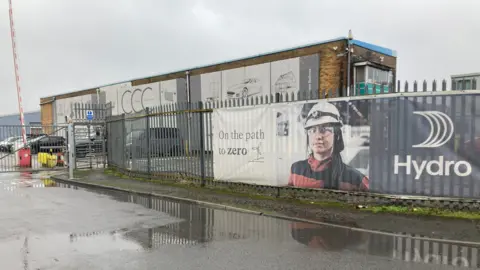 BBC A photo of the Norsk Hydro site in Bedwas, Caerphilly. In the background is a brick building with a metal fence in the foreground. On the fence is a poster with the word Hydro written on it and a picture of a female worker wearing work uniform and a helmet.
