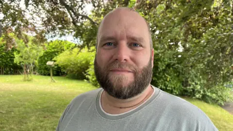 Pete is looking into the camera and smiling slightly. He's wearing a light grey t-shirt, and is stood in a green, grassy area under the cover of a large tree. 