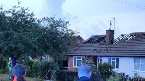 Nuneaton Fire Station A bungalow with a gutted roof near the chimney following a roof fire. The backs of two heads are visible in the foreground and firefighters are standing near a tree
