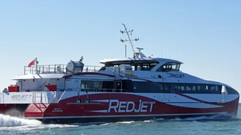 Red and white catamaran travelling on the sea its front it to the right with white spray coming from engines at the rear.