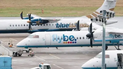 Southampton Airport Two FlyBe commericial aircraft are parked next to each other facing different directions on an airport tarmac. Luggage stairs and ground crew can be seen on the ground, with grass in the background beyond the runway