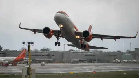 Getty Images An EasyJet aircraft in the air as it leaves the runway at Bristol Airport on an overcast day. Gray hangers can be seen in the background, along with maintenance vehicles and buses. The rear orange tail of an EasyJet plane can be seen to the left of the frame.