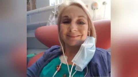 Sue Harley Sue is in a hospital chair with sunlight shining behind her. She has a mask hanging off one side of her face and what appears to be ear plugs in her ears.