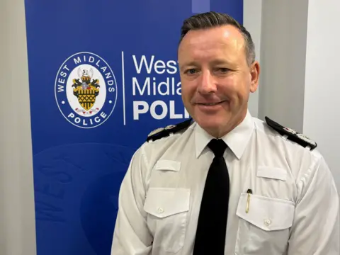 Matt Cooper/PA Media A man with short dark hair, wearing a police uniform smiles at the camera. The sign behind him says 'West Midlands Police'