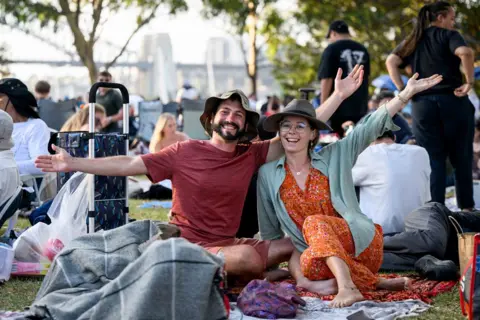 Bianca De Marchi/AAP Image via REUTERS Orang-orang yang bersuka ria berkumpul di Mrs. Macquarie's Point menjelang perayaan Malam Tahun Baru di Sydney, Australia, 31 Desember 2024. Seorang pria dengan kaos coklat dan seorang wanita dengan gaun oranye dan kemeja abu-abu, keduanya memakai topi, duduk di tanah dengan mahkota dan melambai ke arah kamera