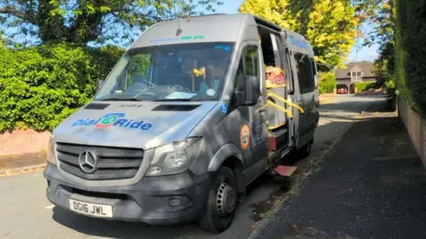 A silver van parked on a residential street. The van is used to operate a community bus service and has the words "Dial a Ride" printed on the bonnet in blue writing. The van's sliding door is open and a female passenger is seated on the bus. A yellow handrail and red steps are extended from the vehicle towards the pavement. 