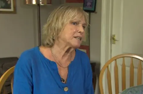 A woman in a blue top looks at the camera as she sits in her kitchen