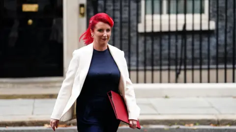 Louise Haigh walking out of 10 Downing Street. She has red hair and is wearing a white blazer with a blue dress. She is also carrying a red folder.