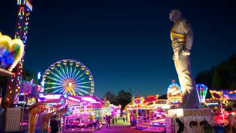Marino Trotta A brightly light fairground at night with a large Michael Jackson statue towering on the right side. His badges and bandolier are painted gold.