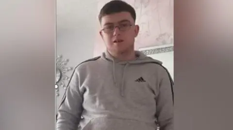 Daniel Ambler, a 23-year-old man with short dark hair, wearing spectacles and a grey hoodie. He is standing in what looks like a living room. There is pink and grey wallpaper on the wall behind him.
