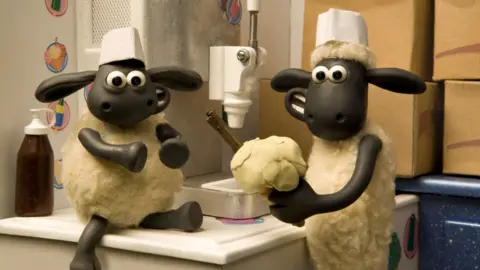 Aardman Two clay animation sheep are in an ice cream van. Timmy is sat ont he counter while Shaun holds a traditional vanilla ice cream with a flake in a cone.
