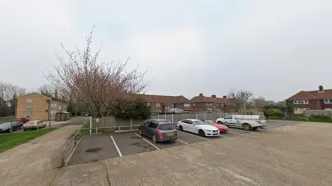 A concrete car park, containing four vehicles, behind a row of modern red brick two-up two-down houses.