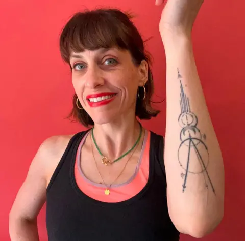 Catherine Hibbert Catherine Hibbert is standing against a red background and wearing a pink and black top. She is showing the camera her geometric shapes tattoo on her left forearm