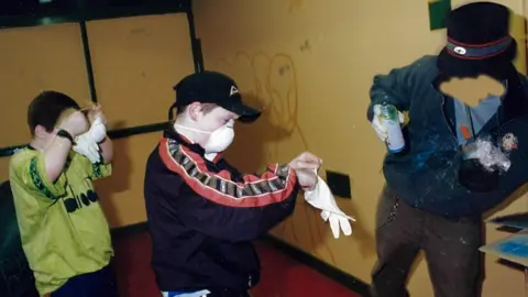 Peter de Boer Banksy leans to one side holding a spray can, while two children put on masks and gloves.