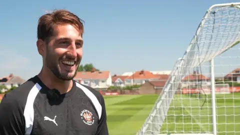 Blackpool goalkeeper Franco Ravizzoli wearing a black Blackpool football team top standing behind the goal at the training ground on a sunny day. He is laughing. 