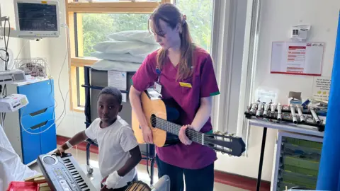Woman with long brown hair in purple top playing a classical guitar with a young boy in a white T-shirt playing a keyboard. They are in a room with white walls and a large window,. There is medical equipment to the left. There is a pipe gamelan on a stand to the right.