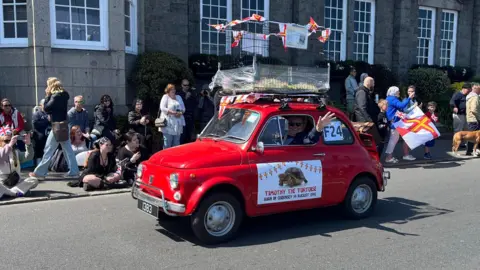 A picture of a red car with the tortoise in a cage on top of it. It has a logo on the side of the car with a picture of the animal. The crowds are lined up along the side of the streets.