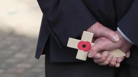 BBC A close up of a man holding a poppy on a wooden cross behind his back. The smartly dressed man is facing away from the camera. He is holding his hands together while holding the poppy.
