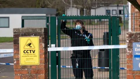 A police officer wearing black uniform and turquoise gloves takes swabs from a green school gate, which is surrounded by blue and white police tape.