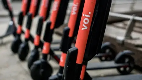 Line of pink and black e-scooters on a pavement - the first is in focus, the others are slightly blurred.