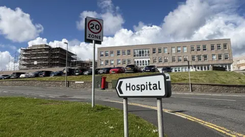 Two grey concrete hospital buildings with windows. The longer one it to the right. To the left, a taller building is covered in scaffolding. Above, a blue sky with clouds. There are signs and a tarmac road in front of the hospital site next to a patch of grass. 