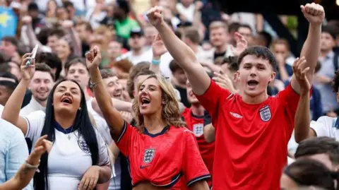 England fans celebrate in a pub garden. A crowd of men and women, in the foreground are two women and two men with their arms up wearing football t-shirts.