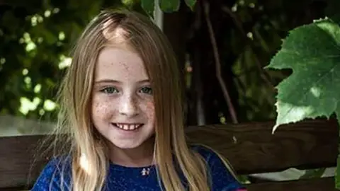 Dale Thomas Cimarron Thomas, a young girl with blonde hair smiling at the camera. She has freckles on her face and is wearing a royal blue top. She is sitting on a bench.